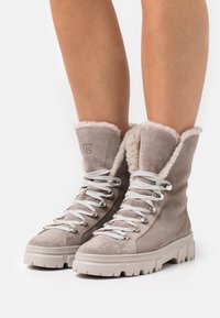 Botas de ante gris con cordones, forradas con piel de oveja blanca, suelas de goma gruesas y detalles metálicos en plateado. Diseñadas para ofrecer comodidad y calidez.