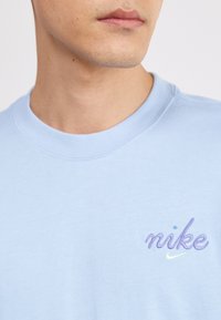 Sweatshirt de algodão azul claro com gola redonda, com o logótipo "nike" em roxo e azul bordado, apresentando uma textura suave e gola canelada.