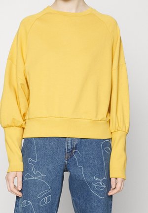Personne portant un sweat-shirt jaune à manches longues et un jean bleu avec des dessins de visages abstraits en lignes blanches sur les jambes.