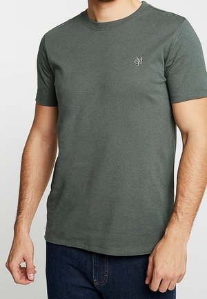 Mand iført en mørkegrøn kortærmet t-shirt med rund hals og lille hvidt logo på venstre bryst, parret med mørkeblå jeans.