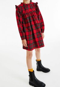 Vestido de tartán rojo con mangas con volantes, cintura fruncida y mangas largas. Combinado con botas negras hasta el tobillo y calcetines amarillos. Fondo blanco.