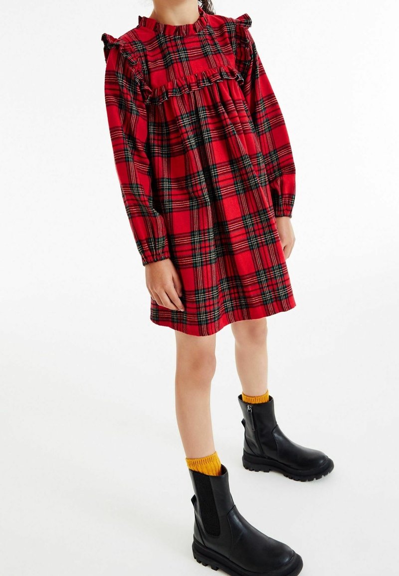 Vestido de tartán rojo con mangas con volantes, cintura fruncida y mangas largas. Combinado con botas negras hasta el tobillo y calcetines amarillos. Fondo blanco.