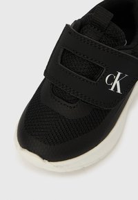 Sapatilha preta com tecido de malha, tira de velcro com o logótipo branco "cK" e sola branca, sobre fundo branco liso.