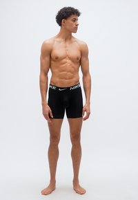 Αθλητικός νεαρός άνδρας με σγουρά μαλλιά, όρθιος ξυπόλητος, φορώντας μαύρα εσώρουχα boxer της Nike, κοιτάζοντας προς τα αριστερά του σε απλό λευκό φόντο.