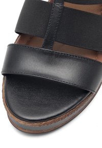 Juoda sandalė su lygiu odiniu viršumi, tampriais dirželiais, apvalia nosimi ir tekstūruota padu, turinčiu rudos spalvos apvadą.