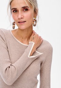 Lichtbruine geribde trui met witte accenten bij de halslijn en manchet. Model draagt grote bewerkte hoepelringen. Handen tonen zichtbare tattoo.