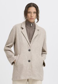 Beige wollen blend jas met reverskraag, knoopsluiting, twee zakken en een relaxte pasvorm. Gedragen over een bruin hoogsluitend top met rits.