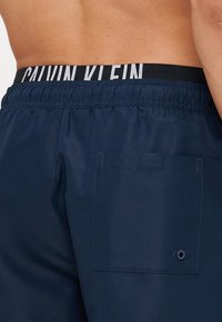 Shorts da nuoto blu con cintura elastica nera caratterizzata dal branding bianco "CALVIN KLEIN". Include una tasca posteriore con dettagli in grommet.
