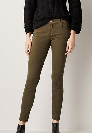 Woman wearing slim-fit olive green pants, black textured sweater, and black ankle boots standing against a plain light background.