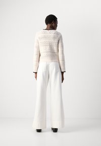 Donna con capelli corti che indossa un maglione lavorato a maglia bianco con motivo, pantaloni bianchi a gamba larga e scarpe nere, in piedi su uno sfondo bianco semplice.