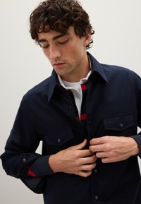 Joven con cabello rizado y oscuro abrochándose una camisa azul marino sobre un polo blanco con rayas rojas y negras, contra un fondo claro y simple.