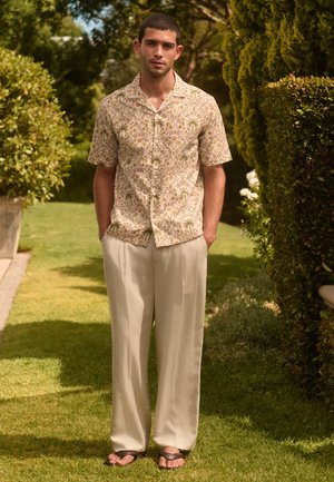 Jeune homme portant une chemise à manches courtes à motifs, un pantalon beige et des sandales, debout sur l'herbe entre des buissons taillés dans un jardin ensoleillé.