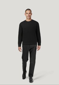 Schwarzer Strickpullover mit einem strukturierten Muster, Rundhalsausschnitt, kombiniert mit dunklen Hosen und schwarzen Schuhen. Model steht vor einem neutralen Hintergrund.