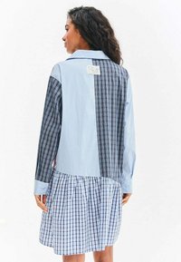 Žena s dlouhými kudrnatými vlasy, která má na sobě volné modré kostkované šaty s více vzory, obrácená zády k jednoduchému bílému pozadí.