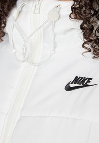 Vit jacka med puffy-textur, med dragkedja och justerbar huva. Svart Nike-logotyp på vänster sida.