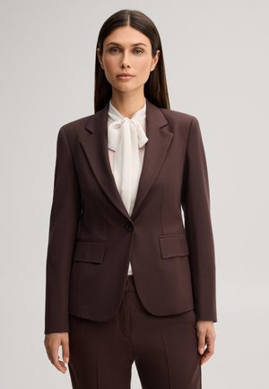 Donna con capelli lunghi e scuri indossa una giacca e pantaloni da completo marrone scuro con una camicetta bianca annodata a fiocco al collo, in piedi davanti a uno sfondo semplice.