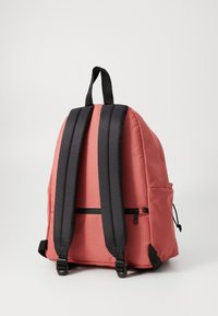 Mochila de tela rosa con correas negras ajustables, un bolsillo frontal con cremallera, bolsillo lateral, forma redondeada y superficie texturizada.