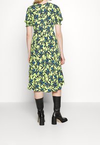 Donna in piedi, indossa un vestito giallo a metà lunghezza con stampa floreale blu e stivali neri con tacco al ginocchio, su uno sfondo semplice e chiaro.