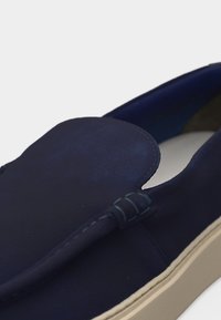 Gros plan sur une chaussure bleu marine sans lacets avec une semelle beige, présentant des détails cousus sur le dessus et une texture lisse en daim.