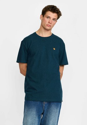 Jonge man met kort bruin haar, gekleed in een donker turquoise t-shirt en blauwe spijkerbroek, staand met handen achter de rug tegen een witte achtergrond.