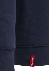 Felpa blu navy con polsini a costine. Presenta un'etichetta rossa con la scritta "hummel" sull'orlo. Tessuto morbido, design minimalista.