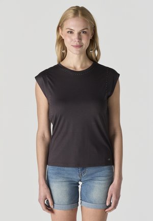 Mujer rubia con pendientes de aro, vestida con una camiseta negra sin mangas y shorts de mezclilla enrollados, posando frente a un fondo liso.