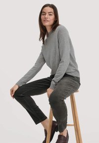 Grijze gebreide trui met lange mouwen en ronde hals; gecombineerd met donkergroene corduroy broek. Model zit op een houten kruk.