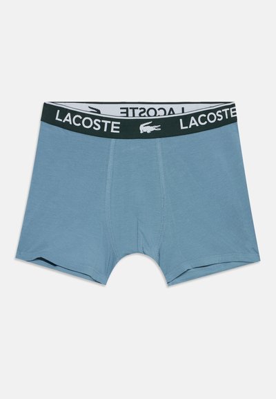 Boxers hommes bleu clair avec ceinture élastique noire affichant le logo blanc Lacoste et l'emblème crocodile.