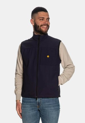 Gilet senza maniche blu navy con colletto alto, zip frontale e tasca sul petto con un accento circolare giallo, abbinato a una camicia leggera a maniche lunghe.