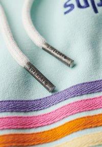 Cordon de sweat à capuche blanc avec embouts métalliques « Superdry » sur tissu bleu clair avec des rayures brodées violet, rose, orange et crème.