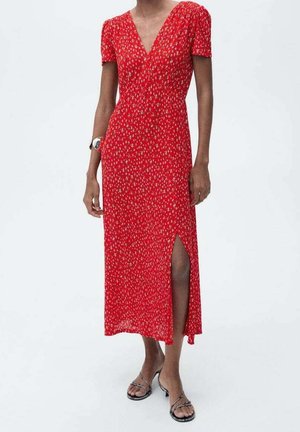 Femme portant une robe midi rouge à manches courtes et col en V, avec un petit imprimé floral et une fente à l’avant de la cuisse, associée à des sandales à talons avec brides.