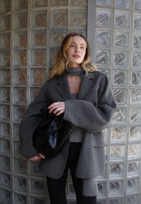 Pilkas per didelis vilnos mišinio švarkas su plačiomis rankovėmis, derinamas su prigludusia pilka palaidine, juodom kelnėm ir juoda odine rankine prieš stiklo blokelių siena.