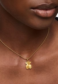 Goldene Halskette mit einem strukturierten, blütenförmigen Anhänger. Die Kette ist mit Perlen versehen und ruht auf dem Schlüsselbein gegen dunkle Haut.