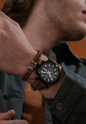 Orologio con quadrante nero e dettagli in argento, cinturino in pelle marrone e particolari cronografici. Abbinato a un bracciale intrecciato marrone con accenti metallici.