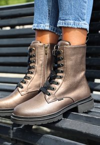Botas de tobillo metálicas en bronce con un acabado texturizado, cordones en la parte delantera, cremallera lateral y cordones negros, que cuentan con una suela de goma negra resistente.