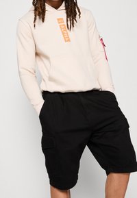 Kremowa bluza z kapturem z logo "ALPHA" w kolorze pomarańczowym, z przednią kieszenią oraz subtelną kieszenią na zamek na rękawie. Czarne szorty cargo z kieszeniami bocznymi.