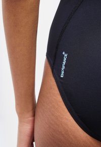 Κοντινό πλάνο ενός ατόμου που φοράει μαύρο μαγιό με μπλε κείμενο «ENDURANCE+» κοντά στο ισχίο, που δείχνει την υφή του δέρματος και τις λεπτομέρειες των ραφών του ρούχου.