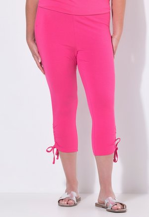 Vrouw draagt felroze cropped leggings met zijstrikjes en zilveren open-toe platte sandalen tegen een effen witte achtergrond.