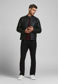 Veste en cuir noir avec un col montant, design ajusté et zips ; associée à un pantalon noir et des baskets blanches ; fond neutre.