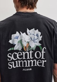 Persona que lleva una camiseta negra con flores blancas y azules y un gran texto que dice "esencia del verano" en la espalda.
