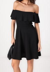 Žena v černých šatech bez ramínek s volánkovým detailem, držící černou kabelku na jednobarevném bílém pozadí.