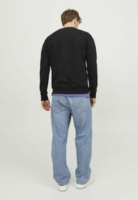 Sudadera negra con cuello redondo acanalado, combinada con unos vaqueros azules holgados. El conjunto se completa con zapatillas blancas, visto desde atrás.