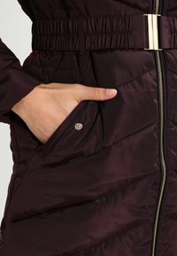 Burgundy pufferjacka med quiltat mönster, elastiskt midjeband och en sidoficka med tryckknappsstängning. Har en framsida med dragkedja och blanka detaljer.