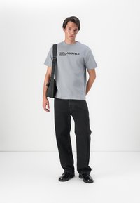 Pelēks kokvilnas T-krekls ar melnu uzrakstu "KARL LAGERFELD JEANS", kombinēts ar melniem džinsiem un melnām kurpēm, pievienota pleca soma.
