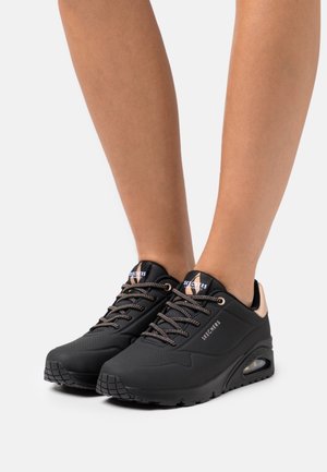 Jambes portant des baskets Skechers noires avec semelles rembourrées, des touches roses et des lacets à motifs sur fond blanc.