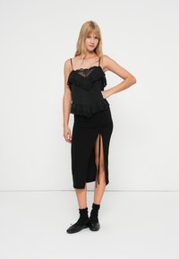 Haut noir à couches avec dentelle, complété par une jupe noire à fente haute. Le mannequin porte des chaussettes noires et des ballerines. Fond minimaliste.
