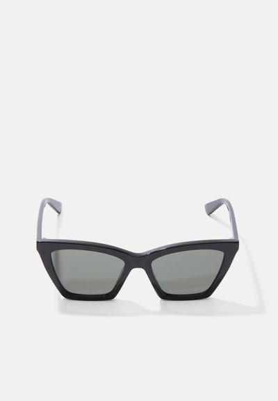 Gafas de sol tipo cat-eye en color negro con lentes tintados en verde, montura de plástico y brazos delgados en gris con acentos plateados. Diseño angular, textura ligera.