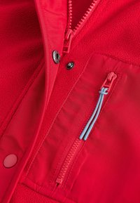 Rode fleece jas met een gestructureerde binnenkant, voorzien van een contrasterende blauwe ritssluiting en drukknoopsluitingen. Ritszak aan de linkerkant.