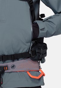 Person iført en grå jakke, der justerer en sort taljerem med en lille pung og et orange sikkerhedsharness bælte.