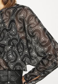 Blouse noire transparente avec motif de pois blancs tourbillonnants. Caractérisée par des bords volantés et des manches larges. Portée avec une ceinture noire texturée.